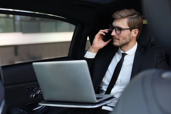 Snygg trygg affärsman i kostym pratar på smart telefon och arbetar med bärbar dator när du sitter i bilen. — Stockfoto