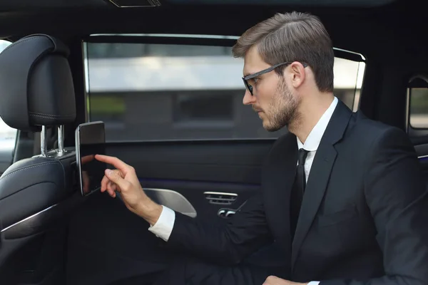 Doordachte jonge zakenman zittend in de luxe auto en het gebruik van zijn Tablet. — Stockfoto