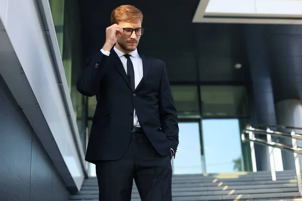 Успешный молодой бизнесмен, держась за очки, спускаясь по лестнице возле офисного здания . — стоковое фото