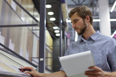 Genç adam ya alışveriş yapıyor ya da bir nalbur deposunda çalışıyor. Tabletindeki malzemeleri kontrol ediyor..