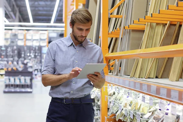 Young man shopping or working in a hardware warehouse standing checking supplies on his tablet. — Stock Photo, Image