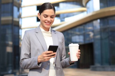 Takım elbiseli güzel bir kadın akıllı telefon kullanıyor ve dışarıda dururken gülümsüyor..