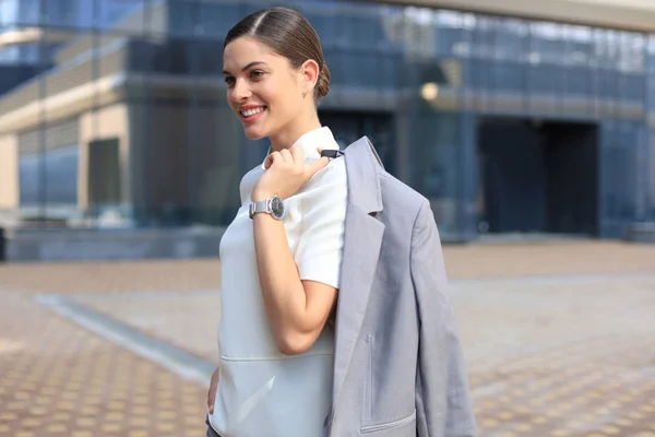 Femme d'affaires souriante debout avec une veste sur son épaule près de l'immeuble de bureaux. — Photo