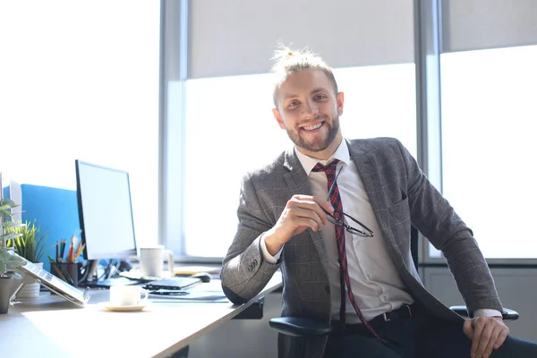 Hezký mladý muž, který se dívá na kameru a usmívá se, když sedí v kanceláři. — Stock fotografie