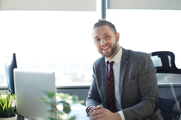 Bello giovane uomo che guarda la fotocamera e sorride mentre è seduto in ufficio . — Foto Stock