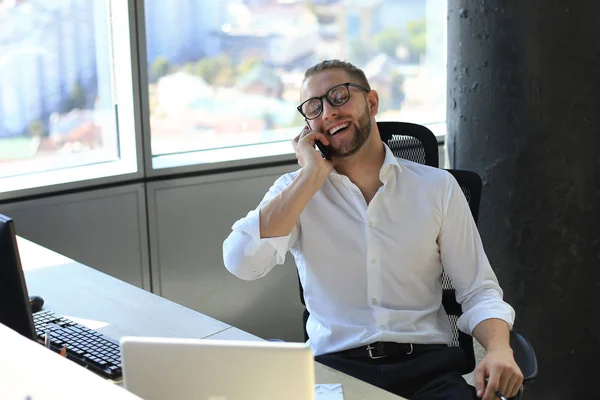 Ωραίος νέος άντρας με μπλούζα που μιλάει στο έξυπνο τηλέφωνο και χαμογελάει ενώ κάθεται στο γραφείο. — Φωτογραφία Αρχείου