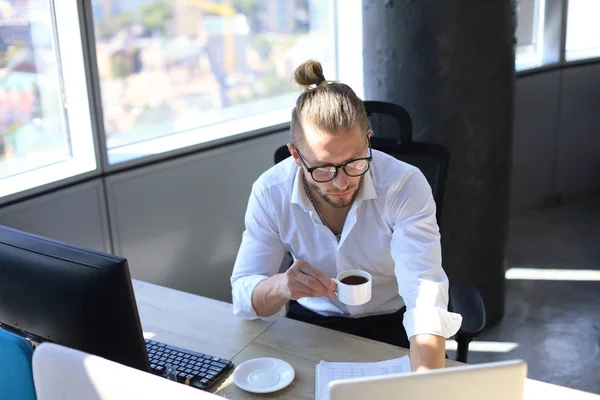Gondolkodó fiatalember inget munka segítségével számítógép és ivás kávét ülve az irodában. — Stock Fotó