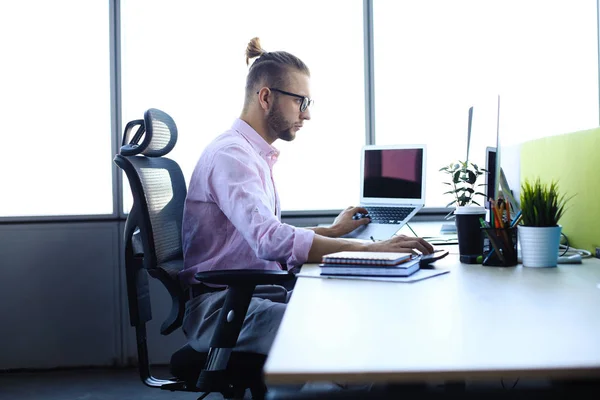 Miło młody człowiek w koszulce pracy przy użyciu komputera, siedząc w biurze. — Zdjęcie stockowe