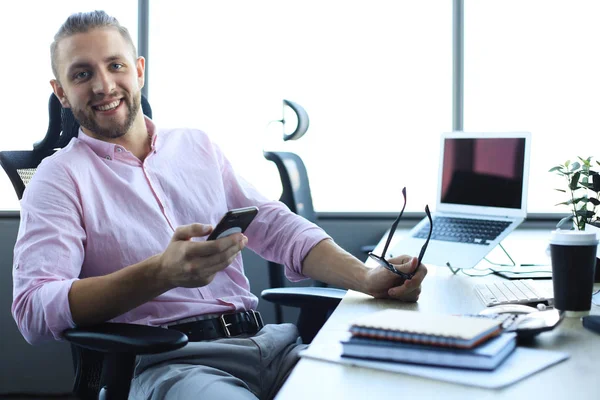 Bom olhar jovem na camisa usando telefone inteligente e sorrindo enquanto sentado no escritório . — Fotografia de Stock