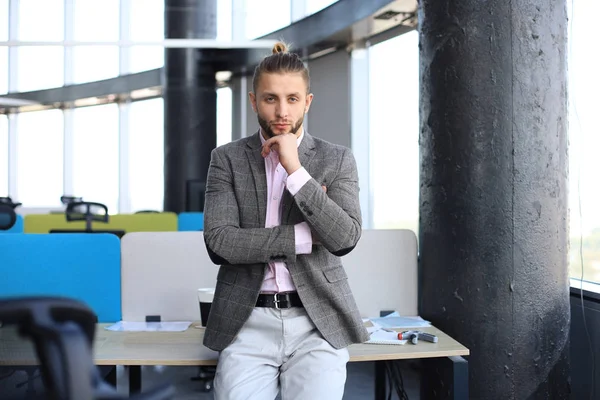 Στοχαστικός νέος επιχειρηματίας σε casual ρούχα κοιτάζοντας την κάμερα και κρατώντας το χέρι στο πηγούνι, ενώ γέρνει στο γραφείο. — Φωτογραφία Αρχείου