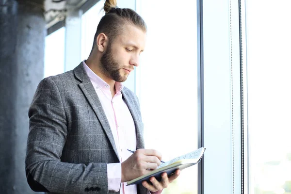 Goed uitziende jonge zakenman in Casual kleding het lezen van zijn notitie pad terwijl staan in het kantoor. — Stockfoto