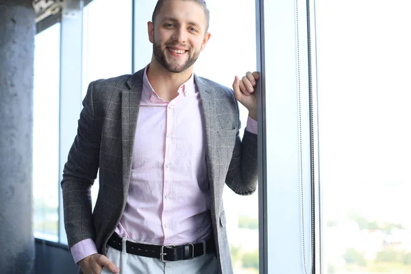 Sonriente joven mirando a la cámara mientras está de pie cerca de la ventana . — Foto de Stock