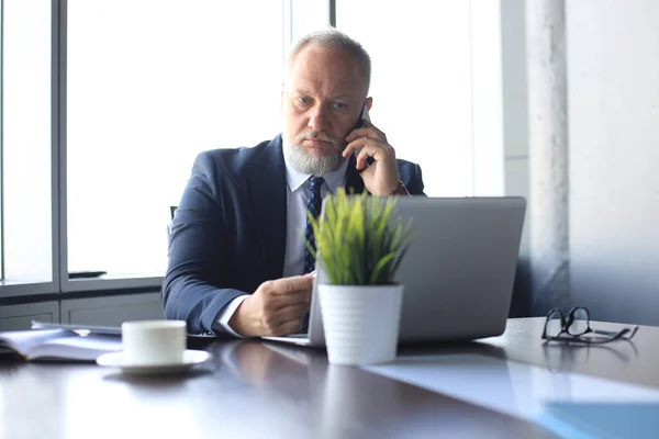 Uomo d'affari maturo che parla sullo smart phone e utilizza il computer mentre lavora in ufficio . — Foto Stock