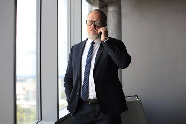 Uomo d'affari maturo che parla su un telefono cellulare vicino alla finestra con vista sulla città. — Foto Stock