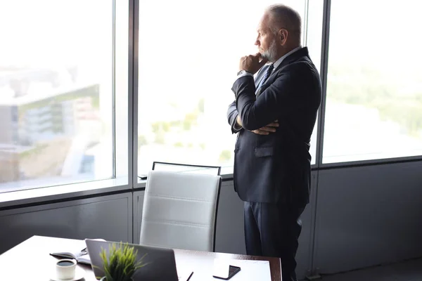 Volwassen zakenman in een corporate Suit staande in kantoor en kijk weg door grote ramen optimistisch. — Stockfoto