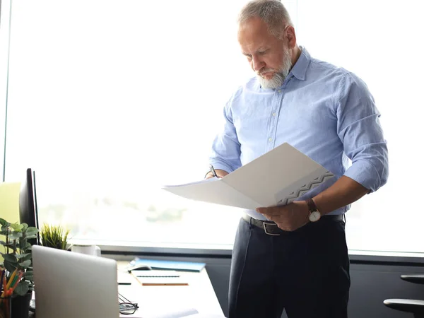 Ernsthafter reifer Geschäftsmann im blauen Hemd liest Vertrag, während er im Büro steht. — Stockfoto