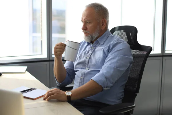 Fokuserad mogen affärsman djupt i tanken när du sitter vid skrivbordet med kopp kaffe i handen i moderna kontor. — Stockfoto