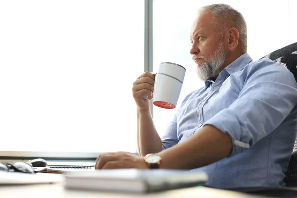 現代のオフィスでコーヒーを片手に机に座りながら、思考に深く焦点を当てた成熟したビジネスマン. — ストック写真