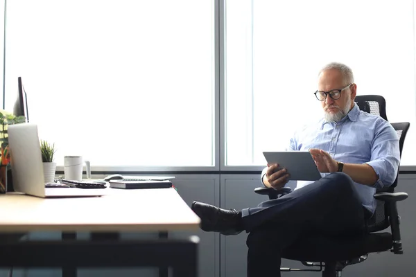 Seriös mogen finansiell rådgivare sitter vid bordet och kontrol lera ekonomisk rapport med hjälp av digital tablett på kontoret. — Stockfoto