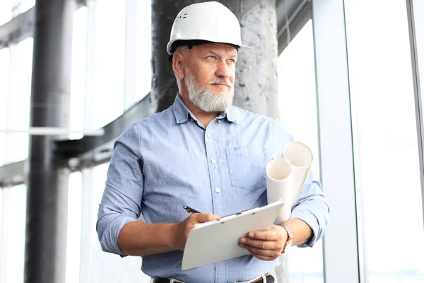 Säker mogen affärsman i HardHat hålla Blueprint och titta bort medan du står inomhus. — Stockfoto