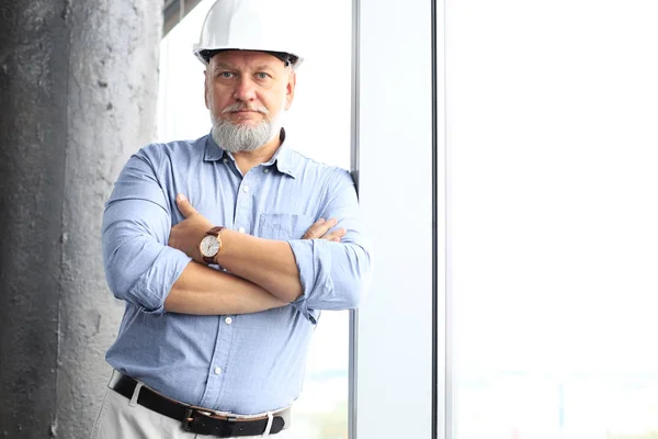 Thoughtful mature architect wearing hardhat in casualwear and looking away while standing near the window in new modern building. — Stock Photo, Image