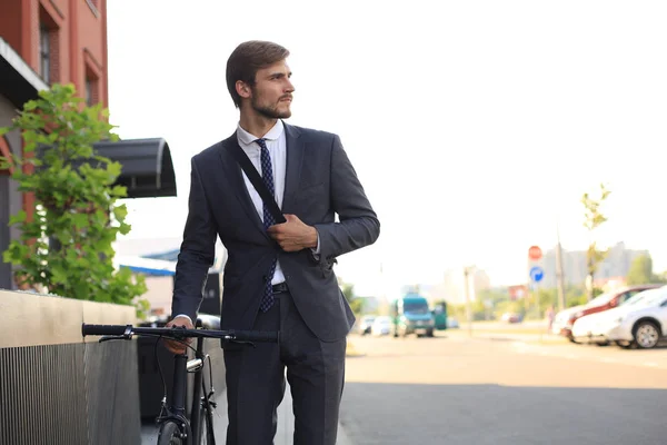 Pensive young businessman walking with bicycle on the street in city.