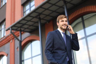Açık havada mobil ayakta konuşurken takım elbise giymiş gülümseyen genç bir iş adamı portresi.