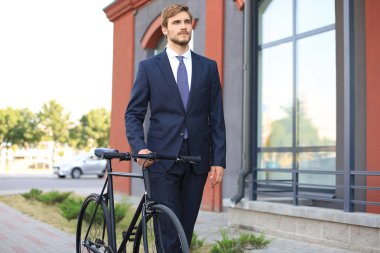 Bir şehir caddesinde bisikletle yürürken takım elbise giymiş genç şık iş adamı.