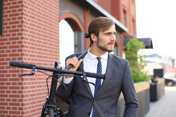 Bonito jovem empresário carregando sua bicicleta no ombro enquanto caminhava ao ar livre . — Fotografia de Stock