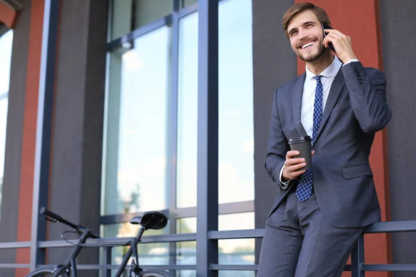 Sonriendo joven hombre con estilo utilizando el teléfono móvil mientras bebe café al aire libre con bicicleta . —  Fotos de Stock