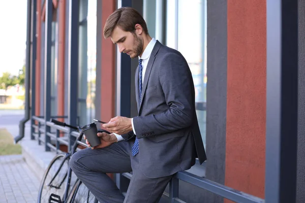 Souriant jeune homme élégant en utilisant un téléphone portable tout en buvant du café à l'extérieur avec vélo . — Photo
