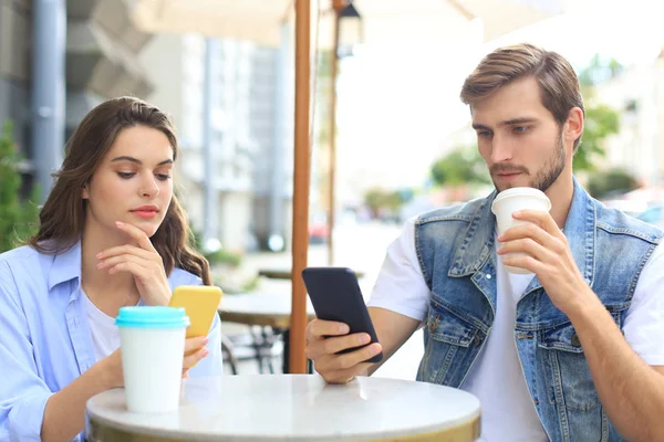Vonzó fiatal pár szerelmes iszik kávét, miközben ül a kávézó asztal a szabadban, a mobiltelefon. — Stock Fotó