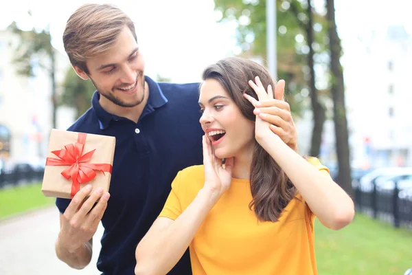 Astonished excited couple in summer clothes smiling and holding present box together while standing on city street. — Stock Photo, Image