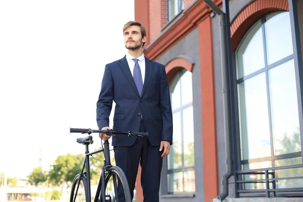 Jovem homem de negócios elegante vestido de terno andando com uma bicicleta em uma rua da cidade . — Fotografia de Stock