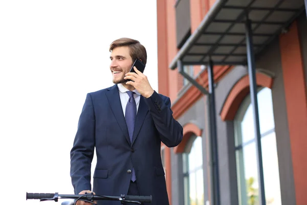Portret van een lachende jonge Business man gekleed in pak praten op mobiel terwijl staand met fiets buitenshuis. — Stockfoto