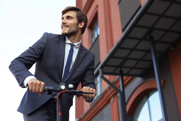 Ung smilende forretningsmand i jakkesæt rider på elektrisk scooter på et forretningsmøde. Økologisk transportkoncept . - Stock-foto