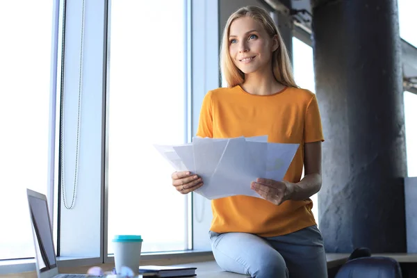 Atractiva mujer de negocios sosteniendo documentos y mirándolos mientras está sentada en el escritorio en la oficina — Foto de Stock