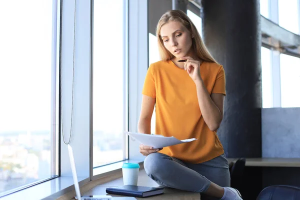 Mulher de negócios atraente segurando documentos e olhando para eles enquanto sentado na mesa no escritório — Fotografia de Stock