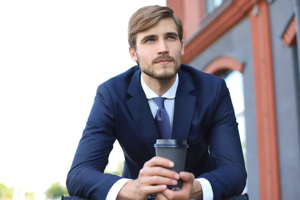 Joven empresario pensativo sosteniendo una taza de café mientras está de pie con su bicicleta cerca del edificio de oficinas en el fondo . — Foto de Stock