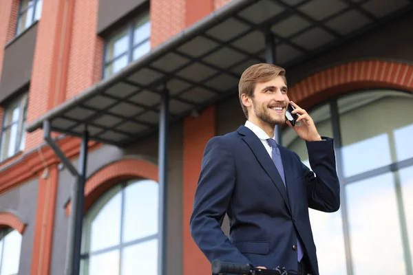 Portré egy mosolygó fiatal üzletember öltözött öltönyt beszél a mobil álló szabadban. — Stock Fotó