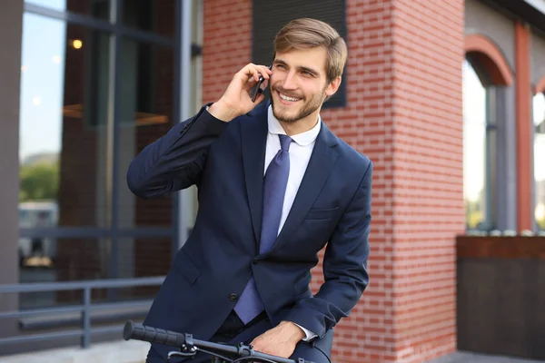 Портрет усміхненого молодого бізнесмена, одягненого в костюм, який говорить на мобільному телефоні, стоячи з велосипедом на відкритому повітрі . — стокове фото