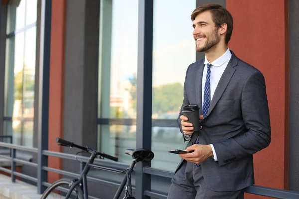 オフィスビルの近くに自転車を持ちながらコーヒーを飲みながら、若いビジネスマンを背景に. — ストック写真