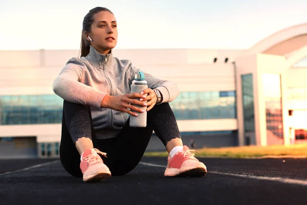 Beautiful young woman in sports clothing drinking water after sport exercise outdoors in stadium