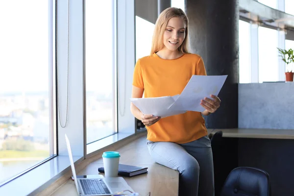 Attractive business woman holding documents and looking at them while sitting on the desk in office