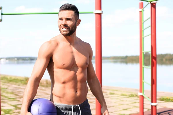 Deportista guapo con torso desnudo descansando mientras hace ejercicio con una pelota de fitness en un día soleado en el parque — Foto de Stock