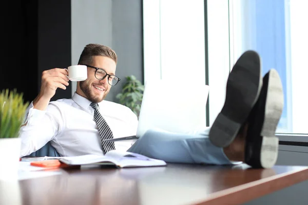 Hombre de negocios guapo sentado con las piernas en la mesa y beber café en la oficina — Foto de Stock