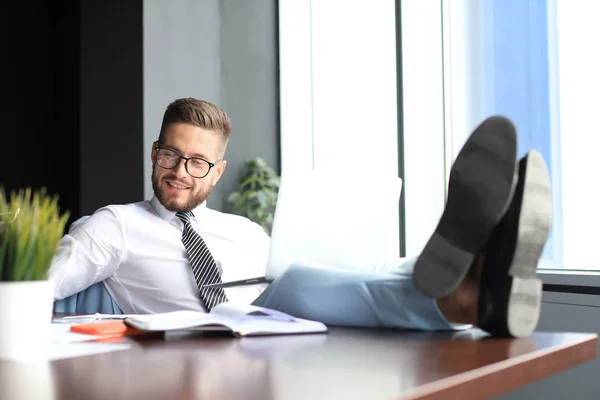 Красивый бизнесмен, сидящий с ногами на столе и использующий ноутбук в офисе — стоковое фото