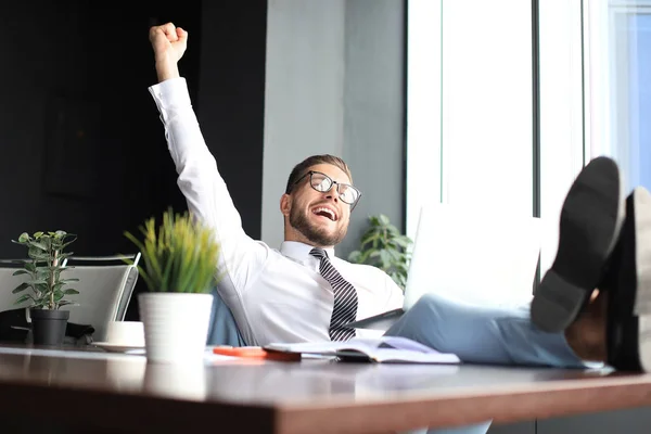 Красивый бизнесмен сидит с ногами на столе, держит руку поднятой и выражает радость в офисе — стоковое фото