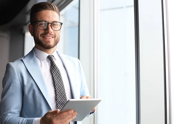 Современный бизнесмен в формальной одежде с помощью цифрового планшета, стоя возле окна в офисе — стоковое фото