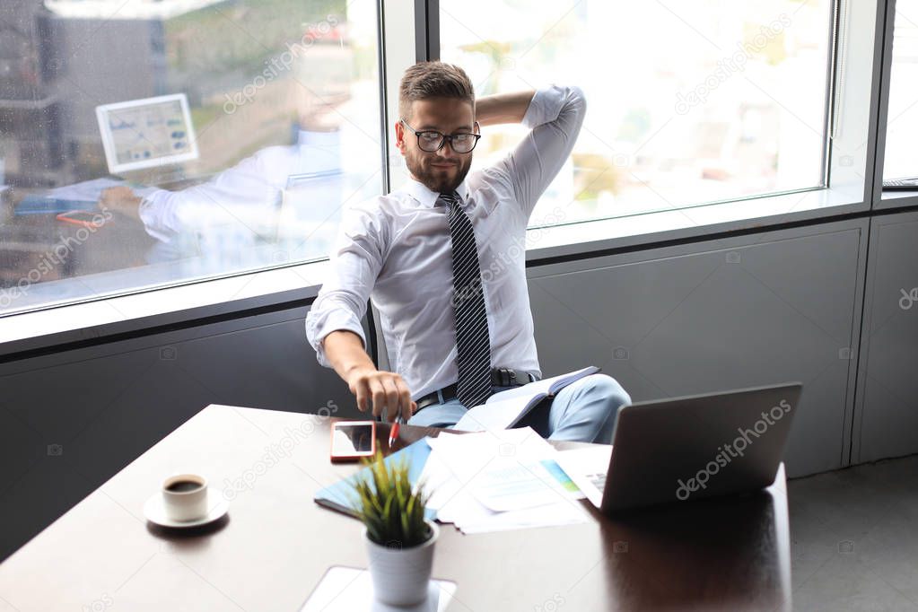 Young modern business man keeping hands behind head and smiling while sitting in the office